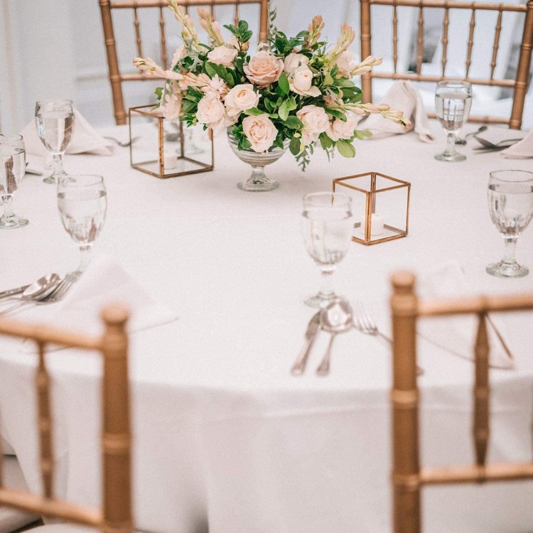 Round table covered with a 90-inch white polyester tablecloth, adorned with a floral centerpiece for elegant dining.
